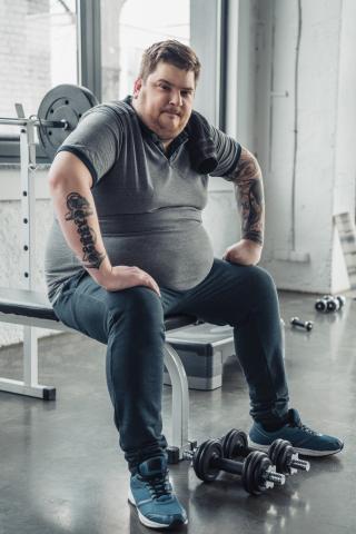Overweight man in gym on weight bench with dumbbells at his feet