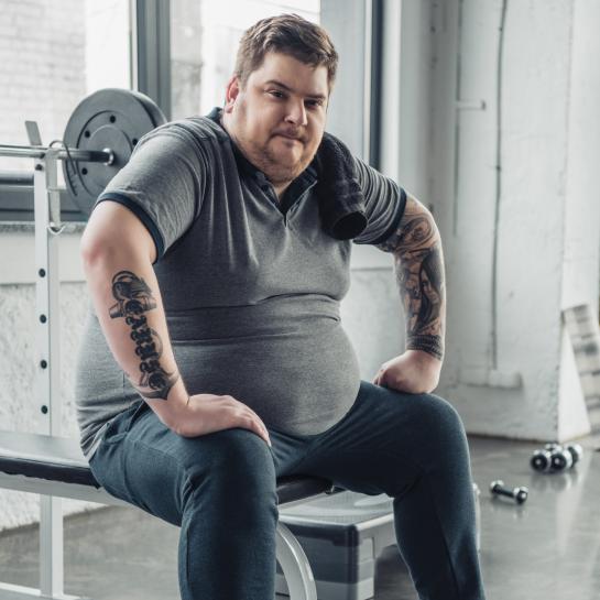 Overweight man in gym on weight bench with dumbbells at his feet